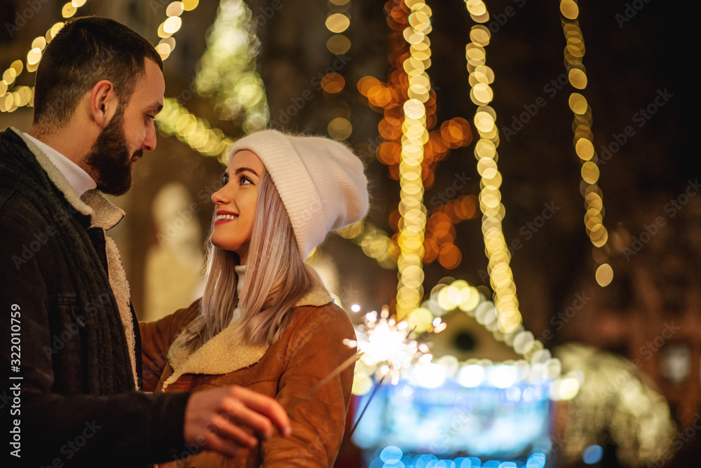 Christmas, winter holidays conception: young happy hugging couple with sparkler, models looking at each other, posing in street of European city. Outdoor night portrait. Copy, empty space for text