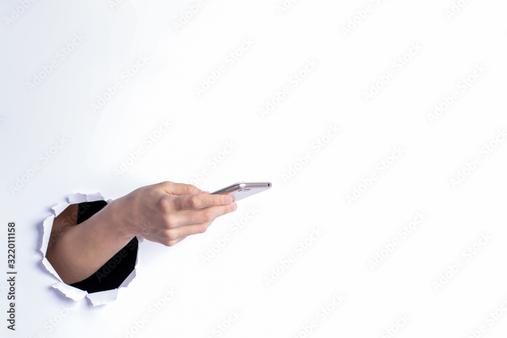 Woman hand holding smartphone through torn hole in white paper wall. Mobile application, online shopping concept. Woman hand holding smartphone through torn hole in white paper wall. Mobile application, online shopping concept.