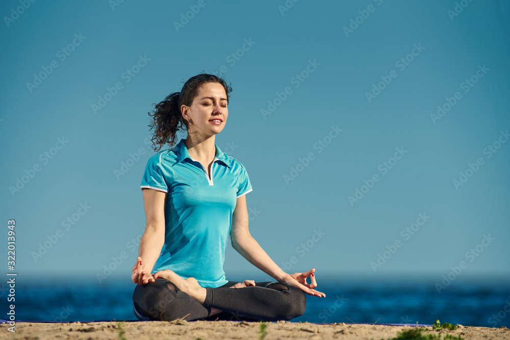 Relaxed young woman meditating while sitting on sandy shore in lotus pose