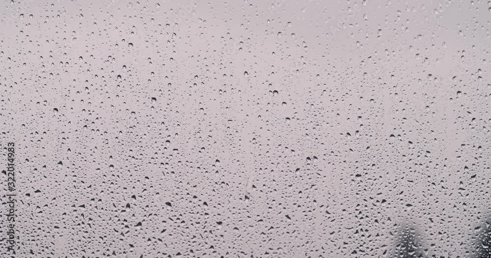 High-contrast raindrops slowly sliding down a window with grey cloudy out of focus background.