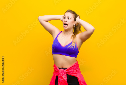 young blonde woman with open mouth, looking horrified and shocked because of a terrible mistake, raising hands to head. sport concept