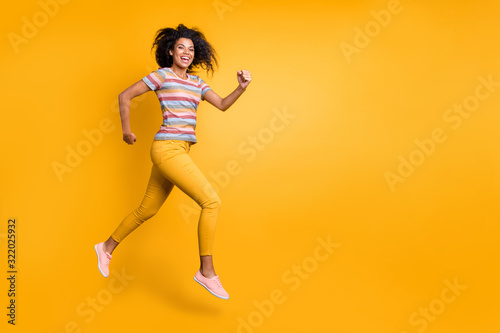 Full length body size view of nice attractive sportive cheerful cheery wavy-haired girl in striped t-shirt jumping running fast flying isolated on bright vivid shine vibrant yellow color background