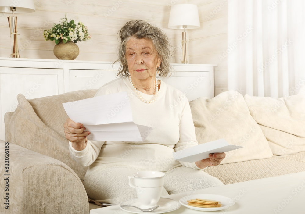 An old woman reads a letter sitting on a sofa - obrazy, fototapety, plakaty 