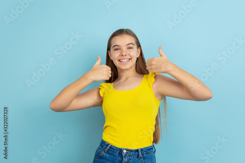Thumbs up. Caucasian teen girl's portrait isolated on blue background. Beautiful model in casual yellow wear. Concept of human emotions, facial expression, sales, ad. Copyspace. Looks cute.
