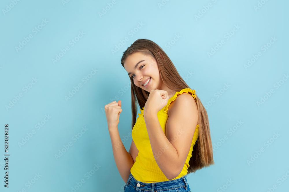 Successful winner. Caucasian teen girl's portrait isolated on blue background. Beautiful model in casual yellow wear. Concept of human emotions, facial expression, sales, ad. Copyspace. Looks cute. Successful winner. Caucasian teen girl's portrait isolated on blue background. Beautiful model in casual yellow wear. Concept of human emotions, facial expression, sales, ad. Copyspace. Looks cute.