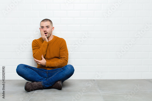 young handsome man open-mouthed in shock and disbelief, with hand on cheek and arm crossed, feeling stupefied and amazed sitting on the floor photo