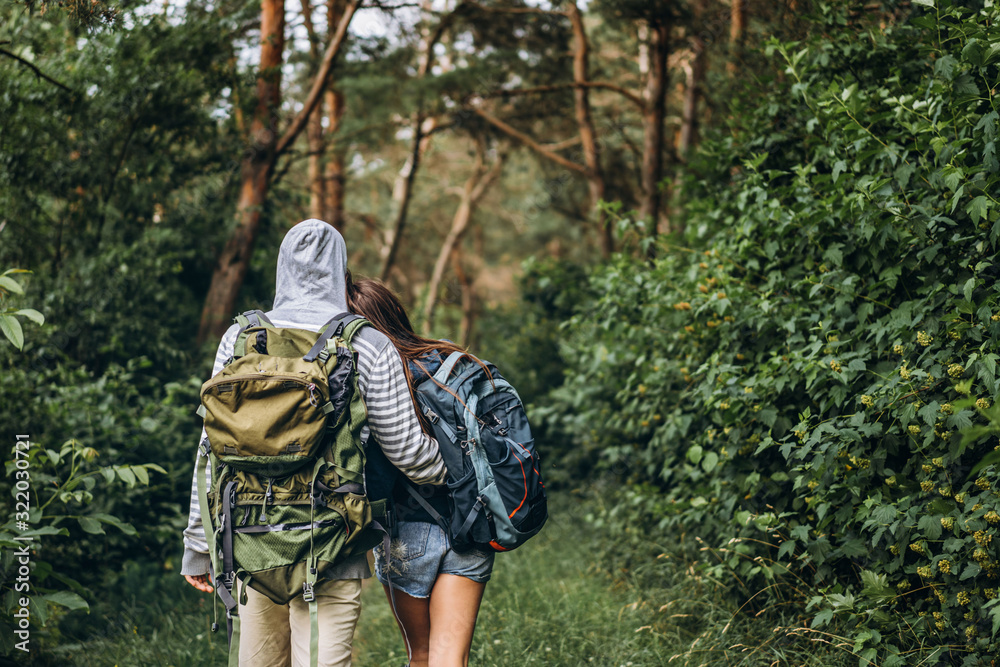 Back view of young couple with backpacks on their backs in the forest. Loving man hugging his beautiful girlfriend on a hike in the woods
