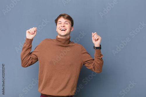 young blonde man looking extremely happy and surprised, celebrating success, shouting and jumping isolated against flat wall photo