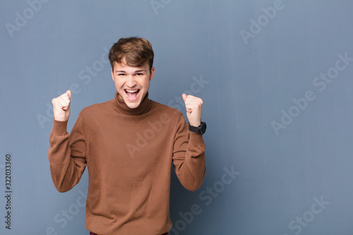 young blonde man feeling happy, positive and successful, celebrating victory, achievements or good luck isolated against flat wall photo