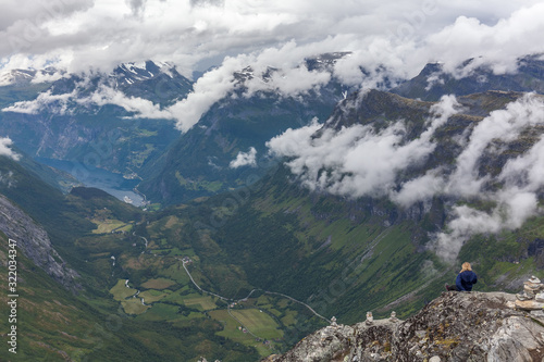 A lonely young girl sits on the edge of a mountain. View to Geiranger fjord and eagle road in cloudy weather from Dalsnibba mountain, serpentine road, Norway, selective focus