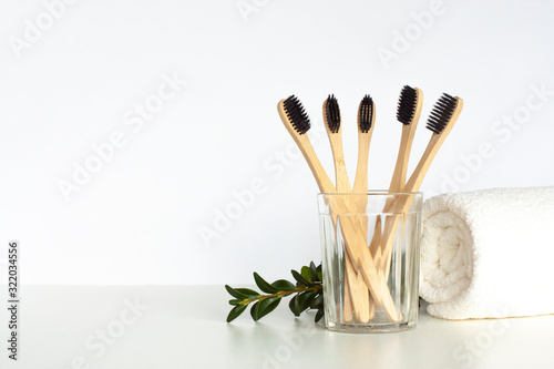 Bamboo toothbrushes stand in glass on light gray table with towel and green twig. Zero waste home, eco friendly bathroom accessories and sustainable lifestyle concept. Copy space.