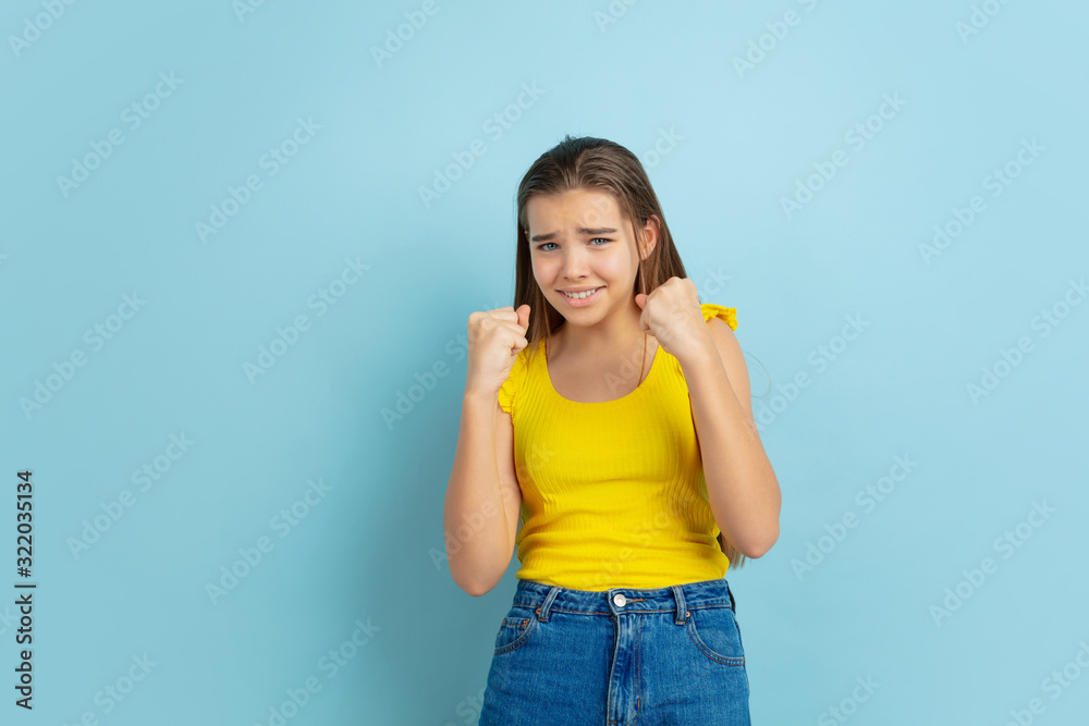 Winning. Caucasian teen girl's portrait isolated on blue background. Beautiful long hair model in casual wear. Concept of human emotions, facial expression, sales, ad. Copyspace. Looks cute. Winning. Caucasian teen girl's portrait isolated on blue background. Beautiful long hair model in casual wear. Concept of human emotions, facial expression, sales, ad. Copyspace. Looks cute.