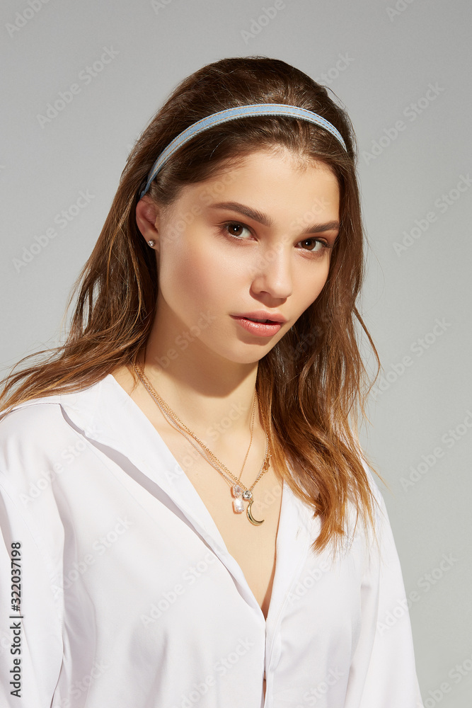 Φωτογραφία A blonde European girl in a white blouse with crystal earrings and golden chains with charms is posing on the gray background