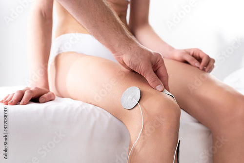 Cropped view of therapist setting electrodes on knee of patient on massage couch on grey background