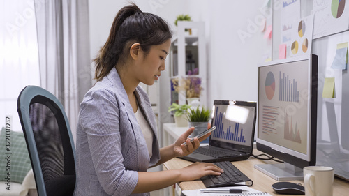 Young elegant asian chinese business woman sitting in home office at table and using smartphone. focus side view beautiful girl employee holding mobile phone checking online website during day.