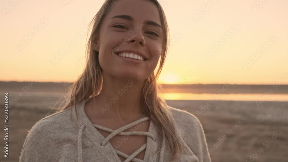 An attractive happy young woman is smiling to the camera while standing near the lake at the dawn time