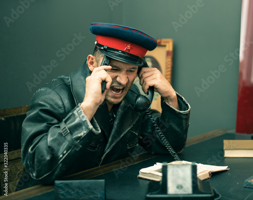 The Soviet man security officer wears cap with red star and hammer and sickle at table in office