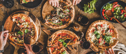 Family or friends having pizza party dinner. Flat-lay of people eating various kinds of Italian pizza and drinking wine over wooden table, top view, wide composition. Fast food lunch concept photo
