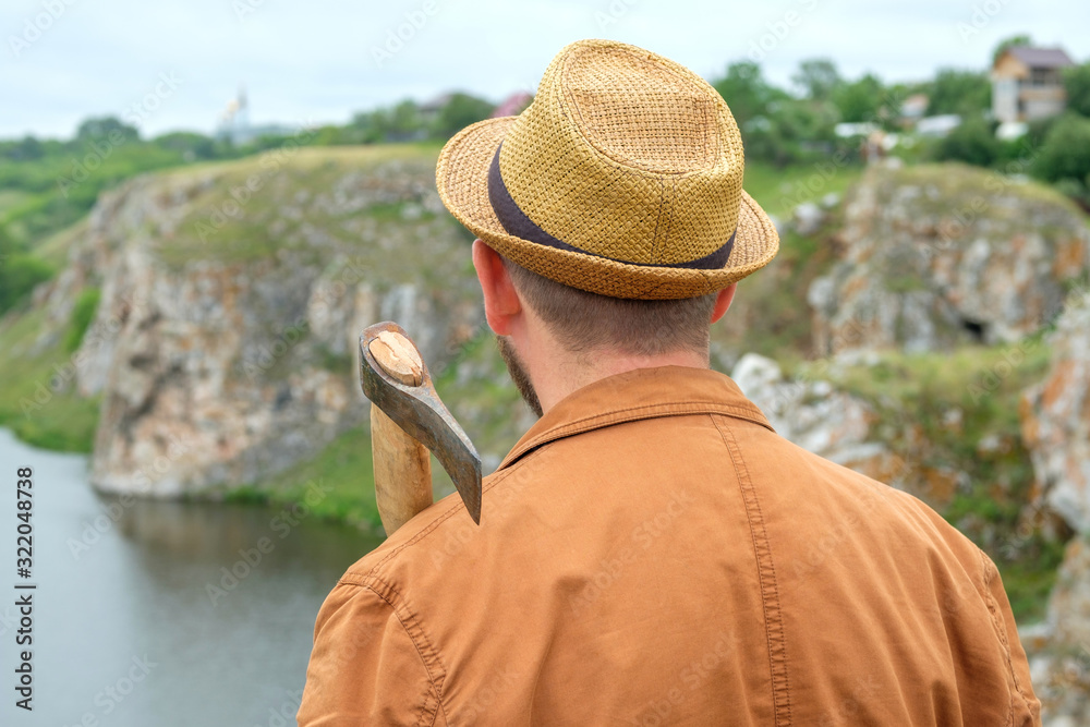 a man in a hat in a brown jacket with an axe on his shoulder background of a rock and a river