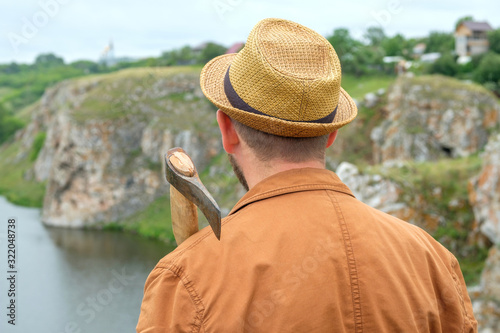 a man in a hat in a brown jacket with an axe on his shoulder background of a rock and a river