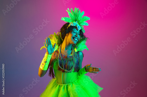 Crazy. Beautiful young woman in carnival, stylish masquerade costume with feathers dancing on gradient background in neon light. Concept of holidays celebration, festive time, dance, party, having fun