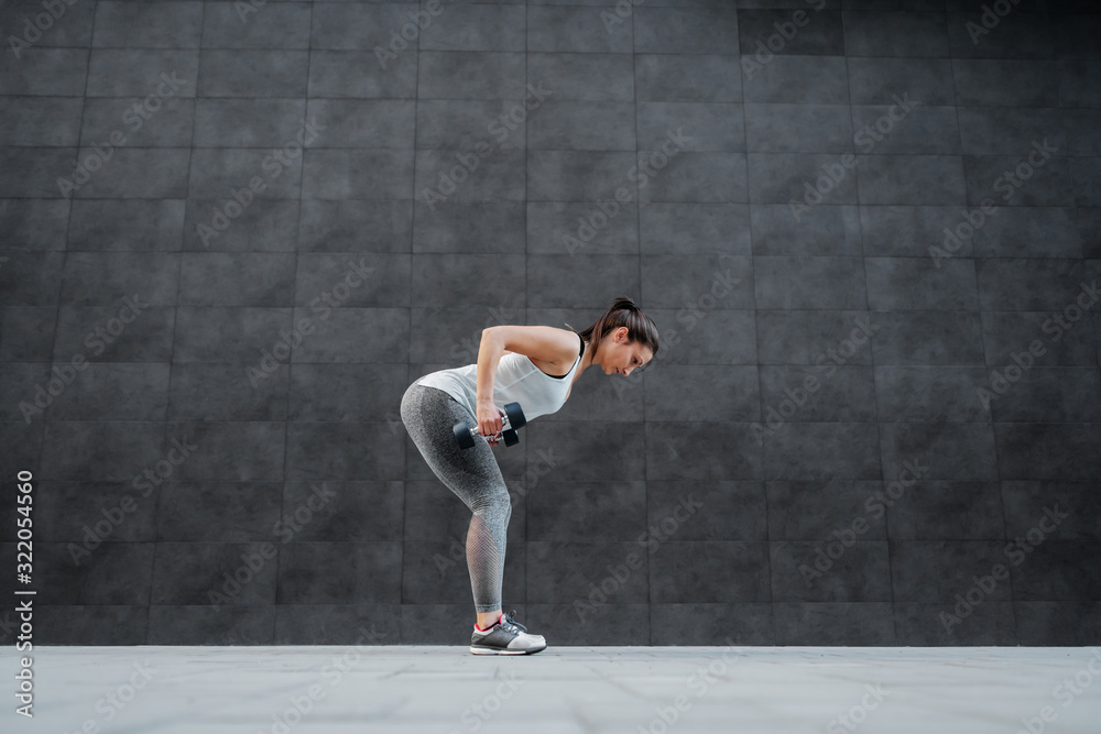 Side view of fit healthy caucasian brunette in sportswear bending and doing exercises with dumbbells in hands. In background is dark wall.