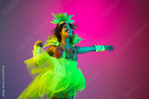 Happiness. Beautiful young woman in carnival, stylish masquerade costume with feathers dancing on gradient background in neon light. Concept of holidays celebration, festive, dance, party, having fun.