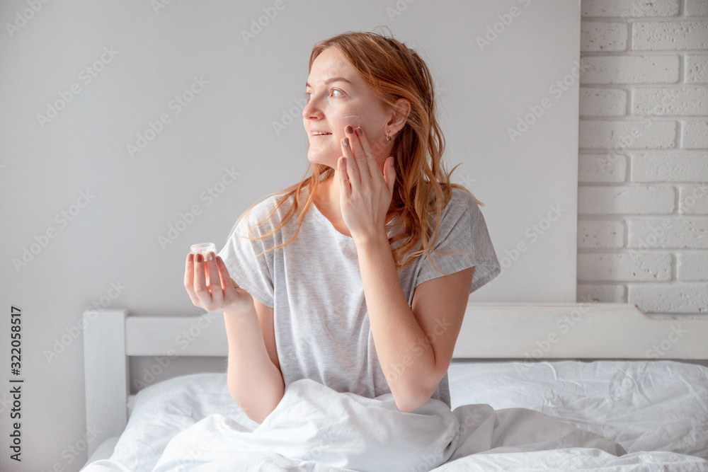 Woman holding bottle of cream with cosmetic products. Blonde woman holding a glass jar with face cream sitting on the bed Woman holding bottle of cream with cosmetic products. Blonde woman holding a glass jar with face cream sitting on the bed