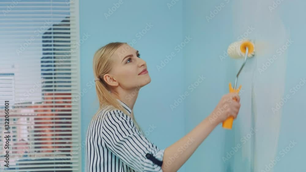 Portrait shot of a young beautiful woman painting a wall at office. She uses white paint and work with a roller. Office renovations. Portrait shot of a young beautiful woman painting a wall at office. She uses white paint and work with a roller. Office renovations.