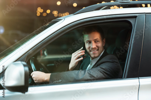 young smiling successful businessman having pleasant phone conversationwhile driving his car with open window, multitasking, big city life, safe driving photo