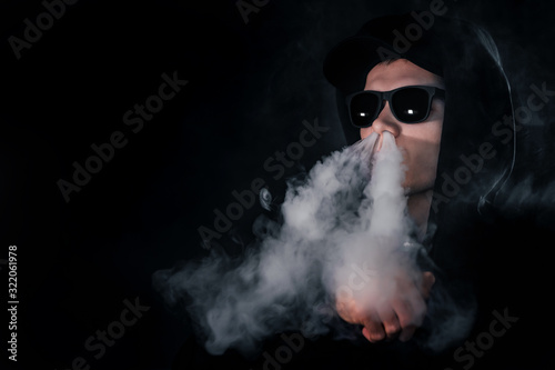 vape man. Portrait of a young white man wearing glasses and a cap lets off steam from an electronic cigarette