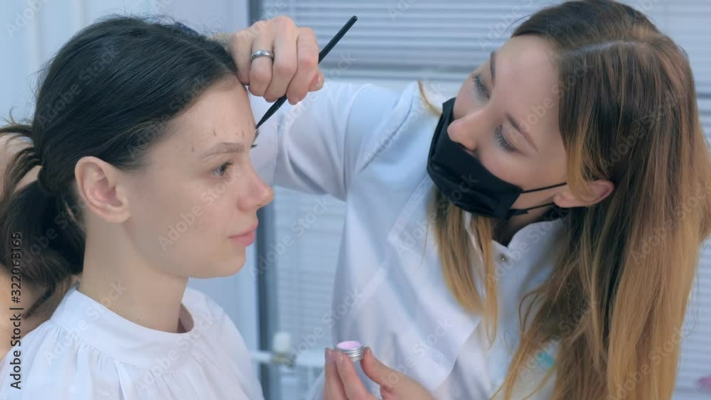Cosmetologist painting brush shape of eyebrows woman before tint procedure in beuty clinic with white paint. Professional beautician at work shaping eyebrows to young girl. Portrait of woman. Cosmetologist painting brush shape of eyebrows woman before tint procedure in beuty clinic with white paint. Professional beautician at work shaping eyebrows to young girl. Portrait of woman.