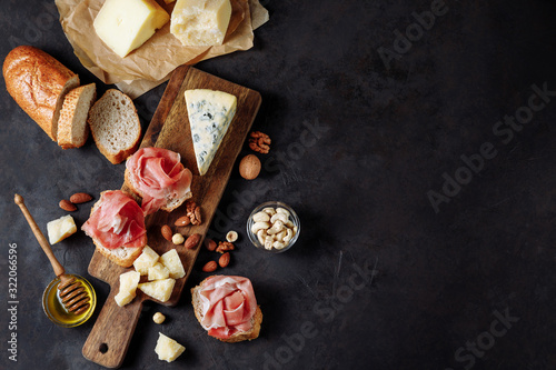 A delicious snack of prosciutto, cheese, nuts and honey. Wooden chopping board. Dark textured background. Appetizer, aperitif, snack table. Top view. Space for text