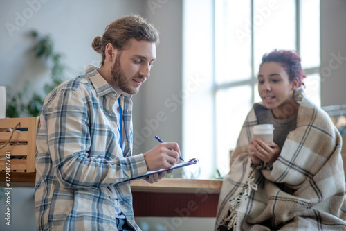 Young man standing writing, girl sitting drinking coffee.