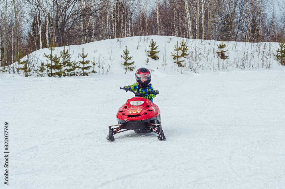 a child in a protective helmet driving a snowmobile rides through a winter meadow in the forest