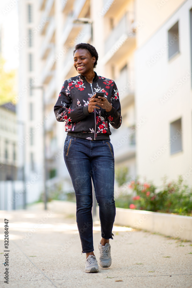 Full body smiling young african woman walking in city with mobile phone