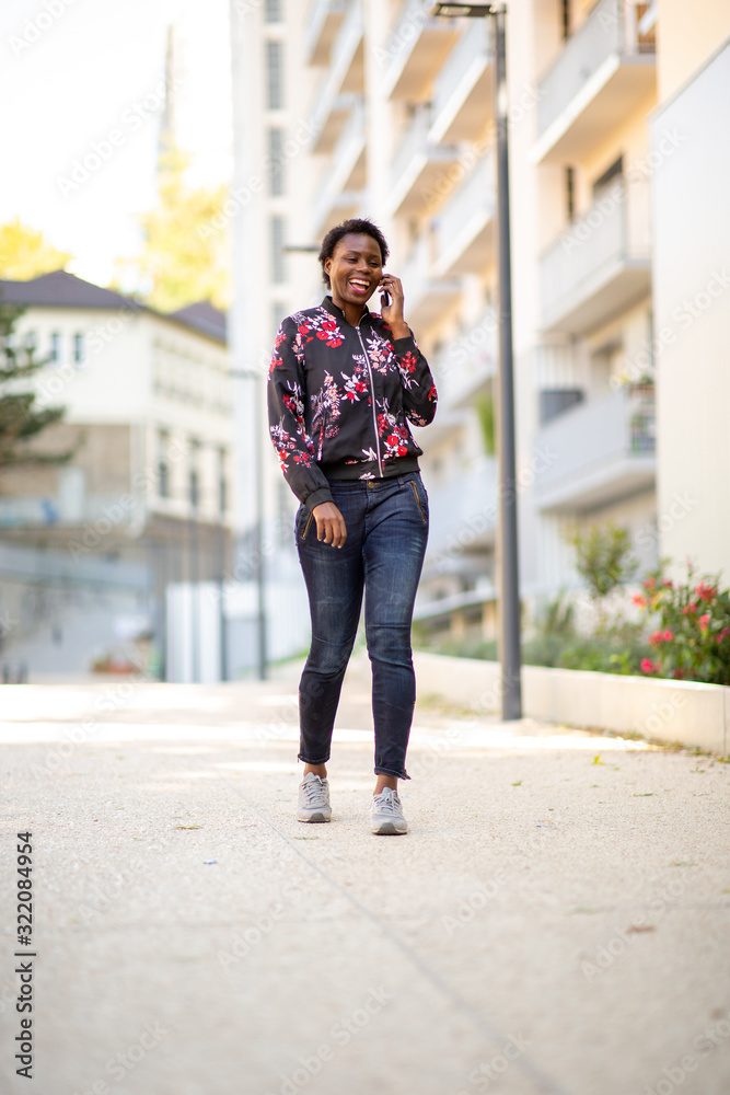 Full body smiling young african woman walking and talking in city with mobile phone