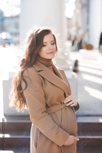 Pretty happy pregnant woman wearing beige jacket posing in street city outdoors. Motherhood. Maternity.