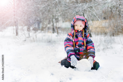 Little beautiful girl sits in the snow and holds a snow globe in her hands. In colored overalls and gloves.