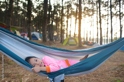 Asian child cute or kid girl sleeping on hammock and happy smiling on nature green tree and pine jungle or forest for summer holiday relax or camping tent picnic and travel trip on family vacation