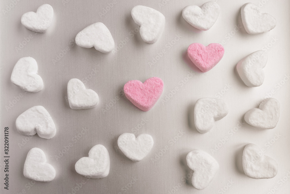 two pink marshmallow hearts lies among white sugar marshmallows in the shape of hearts evenly laid out on a gray aluminum background