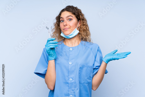 Young blonde woman dentist holding tools isolated on blue background having doubts with confuse face expression