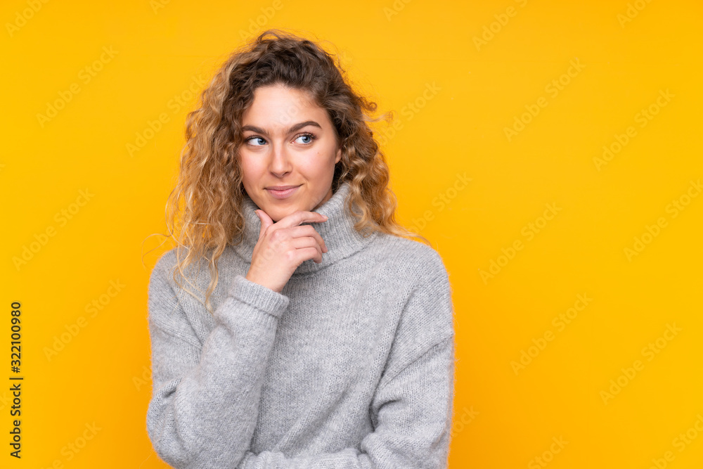 Young blonde woman with curly hair wearing a turtleneck sweater isolated on yellow background looking to the side