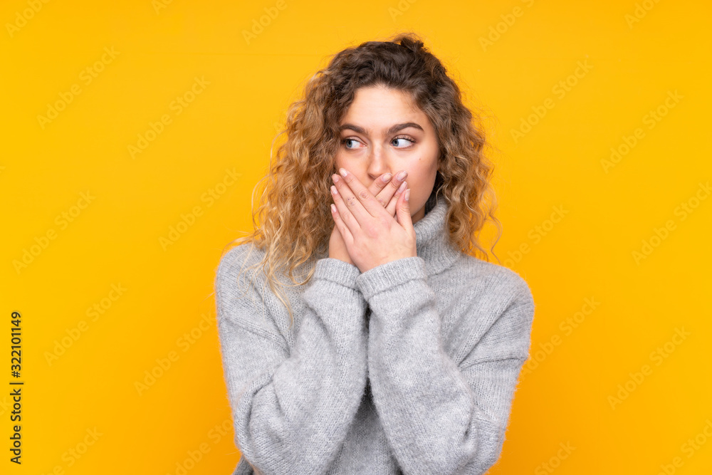 Young blonde woman with curly hair wearing a turtleneck sweater isolated on yellow background covering mouth and looking to the side