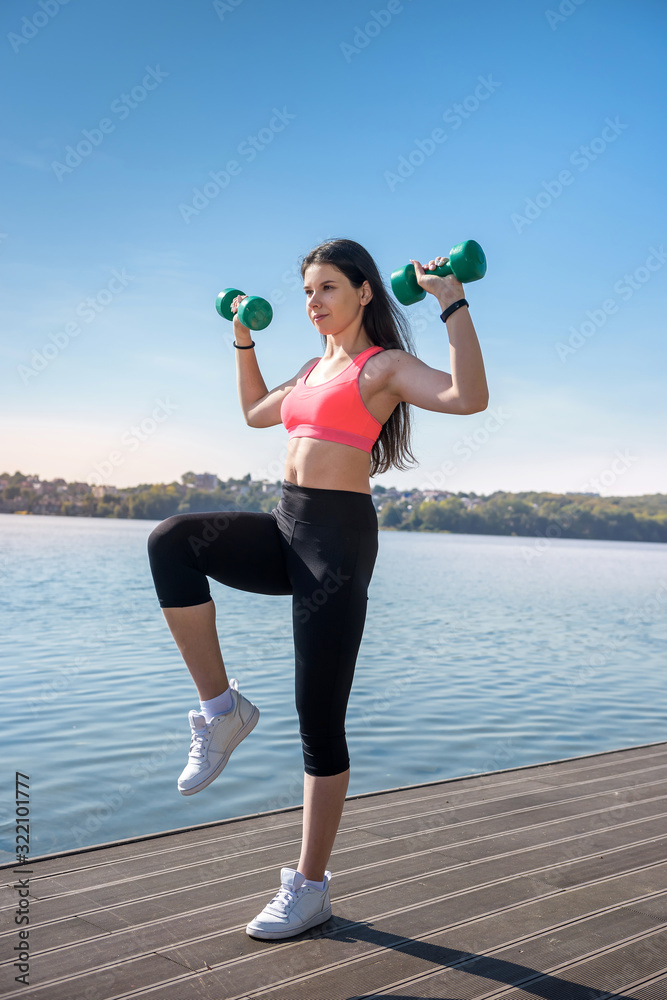 Fit girl doing exercise with dumbbells in her hands at nature, near the lake Fit girl doing exercise with dumbbells in her hands at nature, near the lake