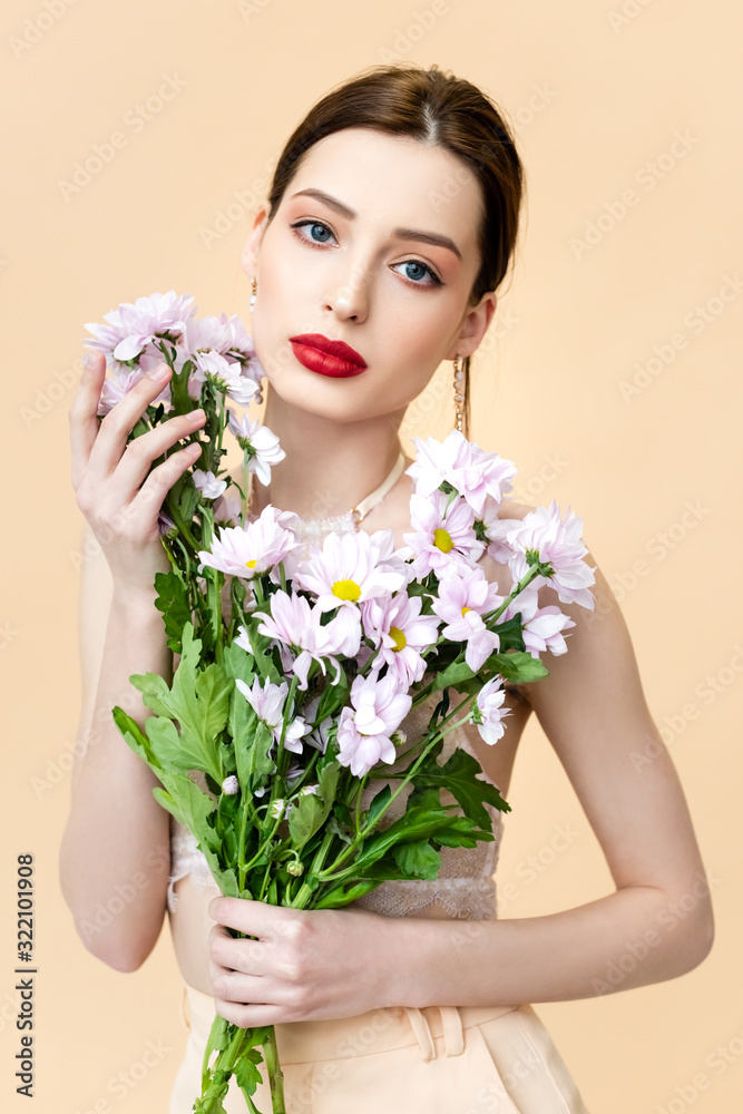 attractive young woman holding blooming flowers and looking at camera isolated on beige