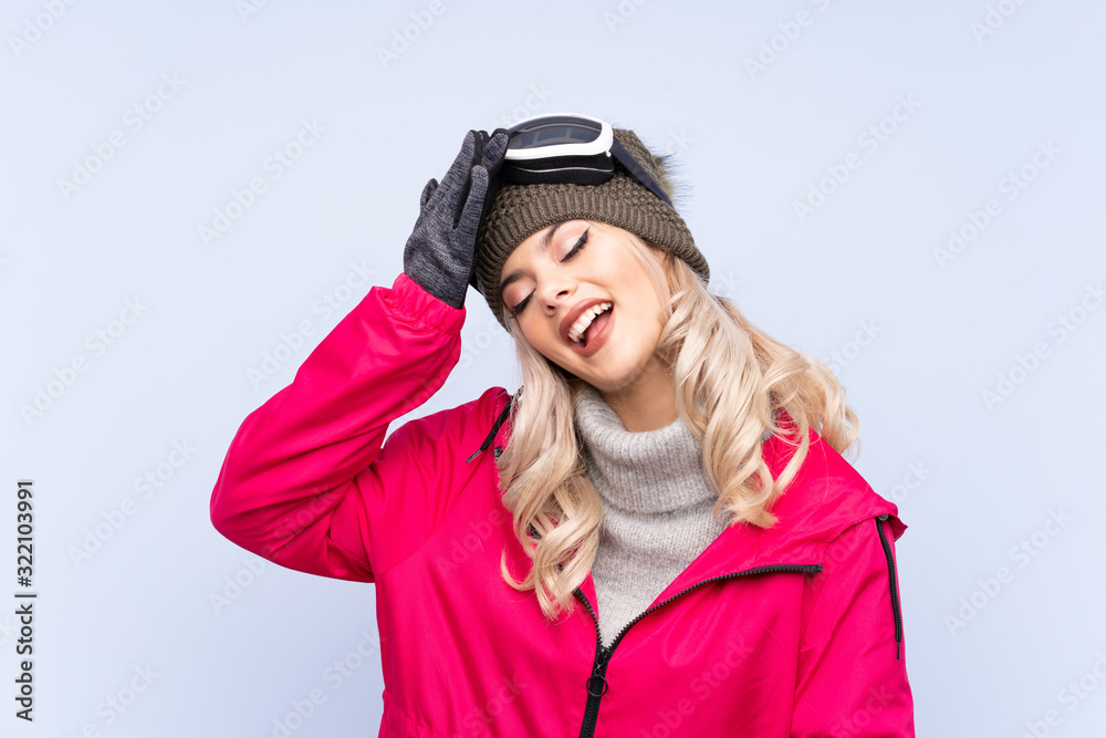 Skier teenager girl with snowboarding glasses over isolated blue background laughing