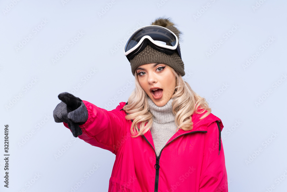 Skier teenager girl with snowboarding glasses over isolated blue background pointing away