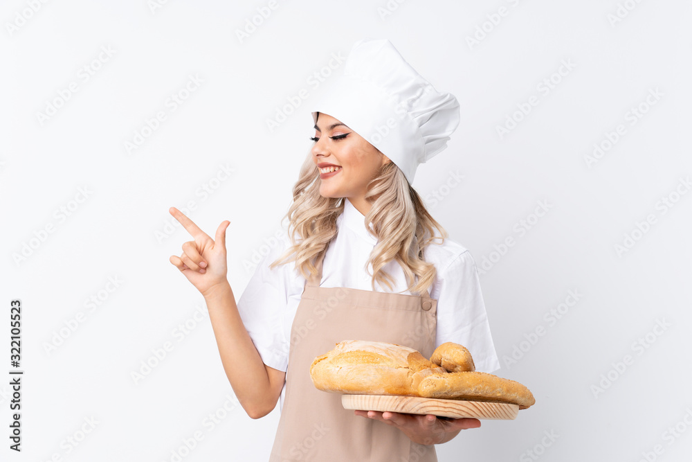 Teenager girl in chef uniform. Female baker holding a table with several breads over isolated white background pointing finger to the side Teenager girl in chef uniform. Female baker holding a table with several breads over isolated white background pointing finger to the side