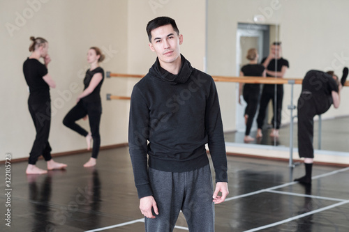Horizontal medium long portrait of young man wearing black and grey outfit standing in rehearsal studio looking at camera